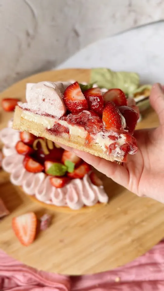 Zemeņu tortes gabals rokā, aizmugurē torte ar rozā krēma rozetēm